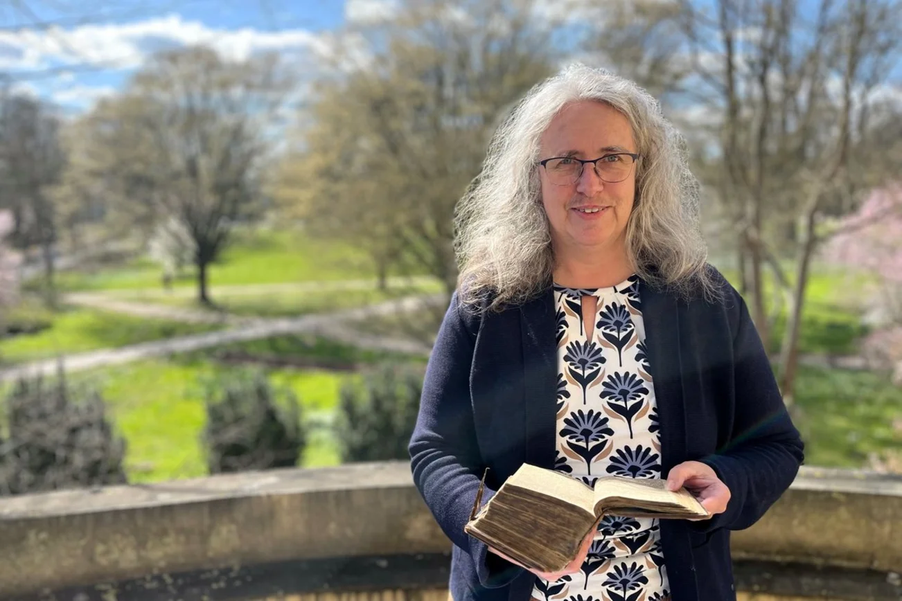 Frau mit historischerm Buch in der Hand steht in Freien mit Park im Hintergrund