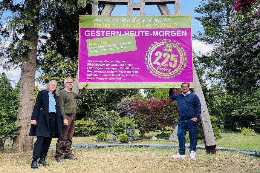 Auf dem Bild stehen Verwaltungsleitung Annett Topaloğlu, der gärtnerische Leiter des Friedhofs, Uwe Pusback, und der Friedhofsbeauftragter im Kirchenkreis Lübeck-Lauenburg, Bernd C. Jakob, vor einem überdimensionalen großen Schild in lila mit der Einladung zum 225. Geburtstags des Lauenburger Friedhofs.  - Copyright: Steffi Niemann