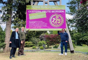 Auf dem Bild stehen Verwaltungsleitung Annett Topaloğlu, der gärtnerische Leiter des Friedhofs, Uwe Pusback, und der Friedhofsbeauftragter im Kirchenkreis Lübeck-Lauenburg, Bernd C. Jakob, vor einem überdimensionalen großen Schild in lila mit der Einladung zum 225. Geburtstags des Lauenburger Friedhofs.  - Copyright: Steffi Niemann