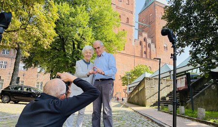 Das Ehepaar Maetzel zeigt mit den Fingern das Sieben-Türme-Symbol, im Vordergrund fotografiert in der Hocke der Fotograf, im Hintergrund ist der Dom zu Lübeck zu sehen