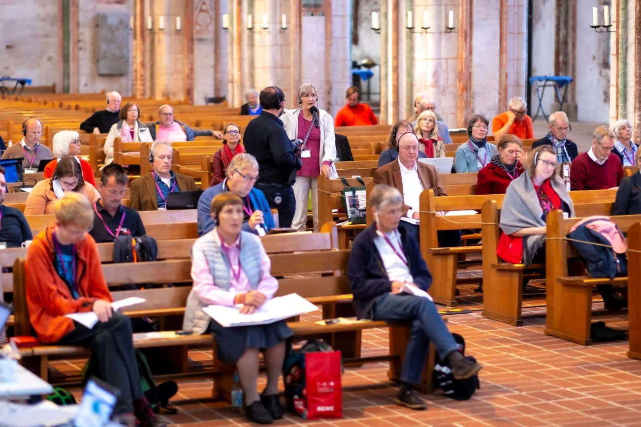 Menschen sitzen in Kirchenbänken, eine Frau steht und spricht in ein Mikrofon. Ein Mann hält das Mikrofon.