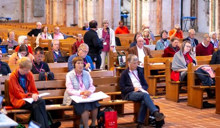 Menschen sitzen in Kirchenbänken, eine Frau steht und spricht in ein Mikrofon. Ein Mann hält das Mikrofon.