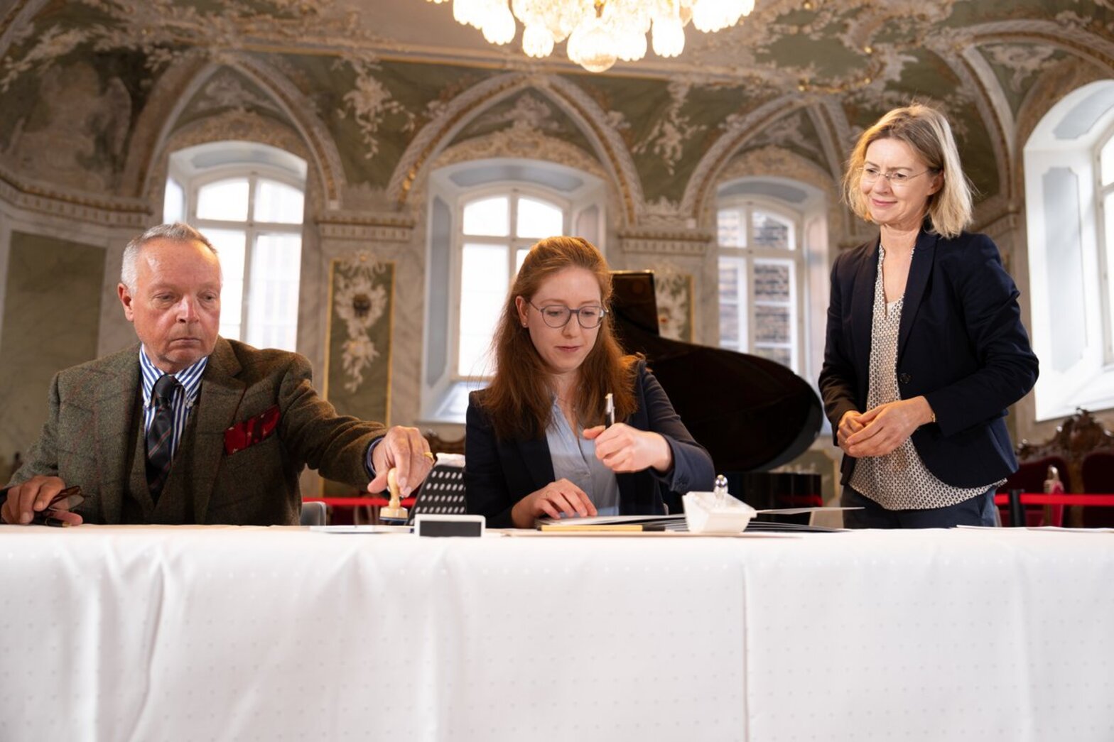 Ein Mann und eine Frau sitzen hinter einem Tisch mit weißer Tischdecke, der Mann ergreift einen Stempel, die Frau rechts neben ihm will gerade ein Dokument unterschreiben, rechts von ihnen steht eine weitere Frau.