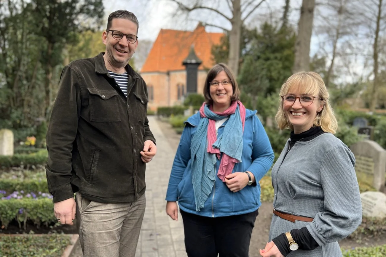 Drei lächelnde Personen auf einem Friedhof