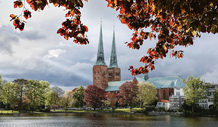 Der Dom ragt mit seinen Domtürmen in den bewölkten Himmel, im Vordergrund ist der Mühlenteich zu sehen, der vom Wind leicht bewegt ist, Am oberen vorderen Rand sieht man rötliche junge Blätter eines Baumes