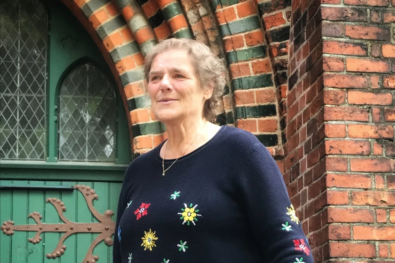 Eine Frau steht vor einem Kircheneingang und lächelt während sie in die Ferne schaut.