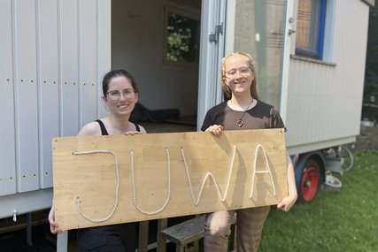 Zwei junge Frauen stehen vor einem Bauwagen und halten ein Schild mit der Aufschrift 'JuWa'. - Copyright: Annkathrin Bornholdt