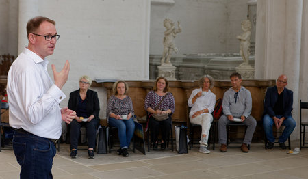 Bürgermeister Jan Lindenau steht im Vordergrund links, ein Teil der Kalenderpaten und Sieben Türme Teammitglieder sitzen auf Holzbänken im Dom zu Lübeck