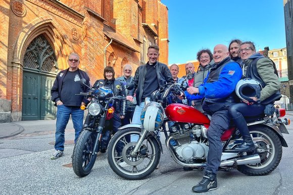 Eine Gruppe Menschen stehen mit zwei Motorrädern vor einer Kirche. 