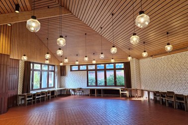 Blick in den großzügigen Gemeindesaal im Standort Düneberg mit Holzvertäfelter Decke und von der Decke herabhängenden Kugelleuchten. Die Fensterfronten an den beiden außenänden lassen viel licht hinein, der Boden ist gefließt. 