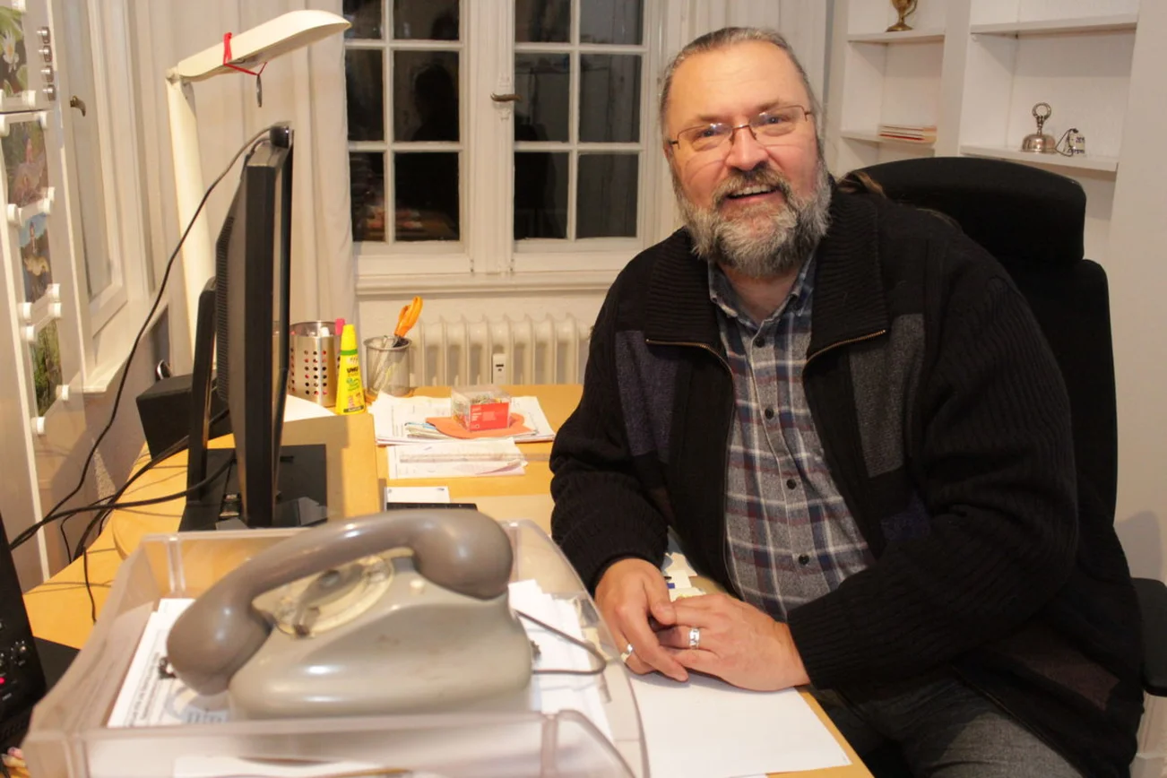 Ein Mann mit grau meliertem Bart, hoher Stirn und unauffälliger Brille sitzt an einem Schreibtisch und lächelt in die Kamera. Im Vordergrund steht in einem Eingangskorb ein graues Wählscheibentelefon.