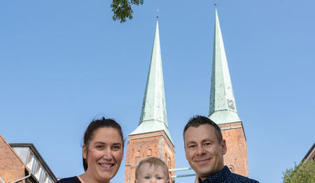 Familie Kosakowski stehen nebeneinander, inder Mitte ihr Kleinkind, im Hintergrund sind die beiden Türme vom Dom zu Lübeck zu sehen. Der Himmel ist strahlend blau und über dem Ehepaar sieht man das grüne Blätterdach eines Baumes