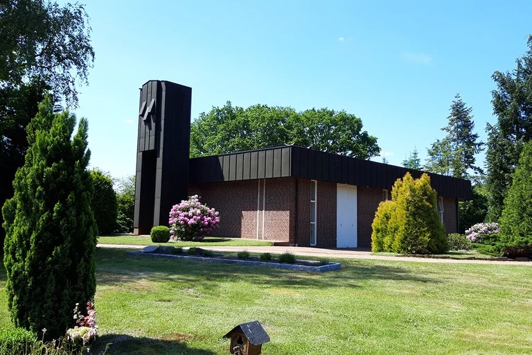Kapelle Waldfriedhof steht im Hintergrund bei strahlend blauem Himmel. Links und rechts sind Bäume und Sträucher. Im Vordergrund Rasen und links ein kleiner grüner Baum