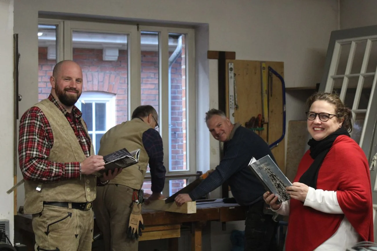 In einer Werkstatt schauen zwei Menschen freundlich in die Kamera. Der Mann links trägt traditionelle Maurerkleidung und die Frau rechts einen roten Poncho. Beide halten ein hellblaues Buch in der Hand. Im Hintergrund arbeiten zwei Männer an einem Balken.