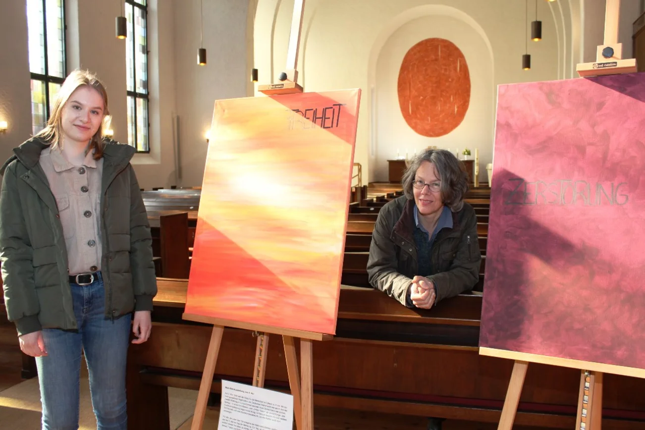 Zwei Frauen stehen zwischen zwei Staffeleien, darauf stehen großformatige Bilder. Im Hintergrund ist der Altarraum der Lutherkirche Lübeck erkennbar.