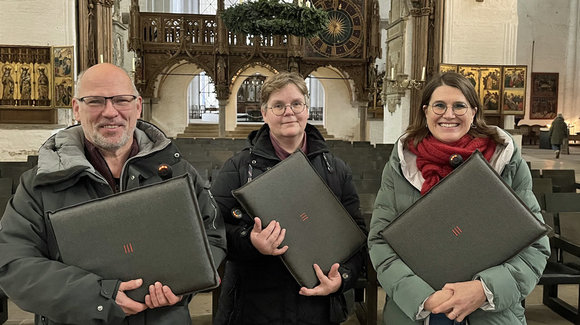 Ein Mann und zwei Frauen stehen in einer Kirch und halten graue Sitzkissen in den Händen