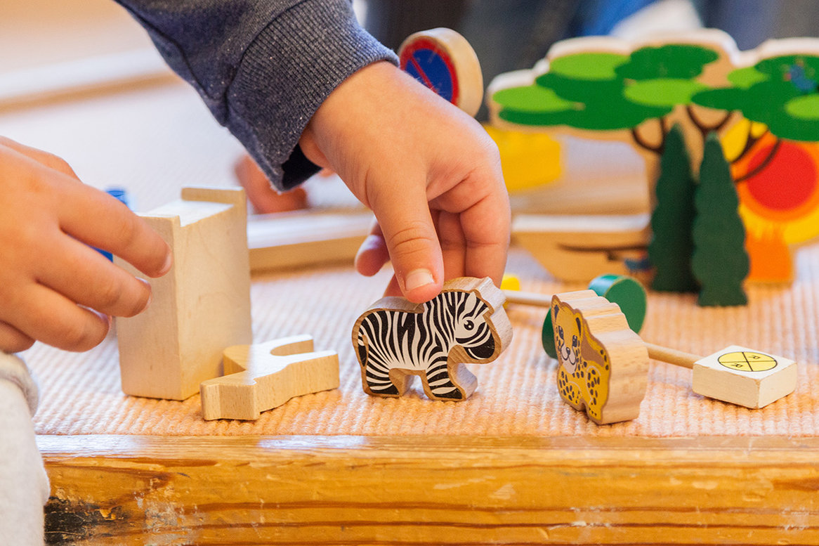 zwei Kinderhände spielen mit Holzfiguren, welche auf einem braunen Holztisch liegen