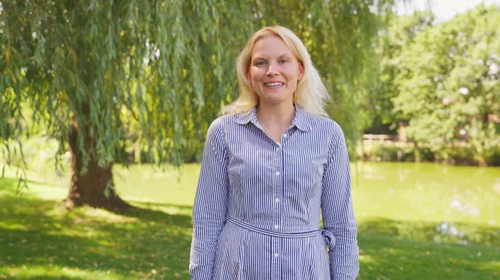 Eine blonde Frau mit gestreiften Kleid steht vor einem Baum.