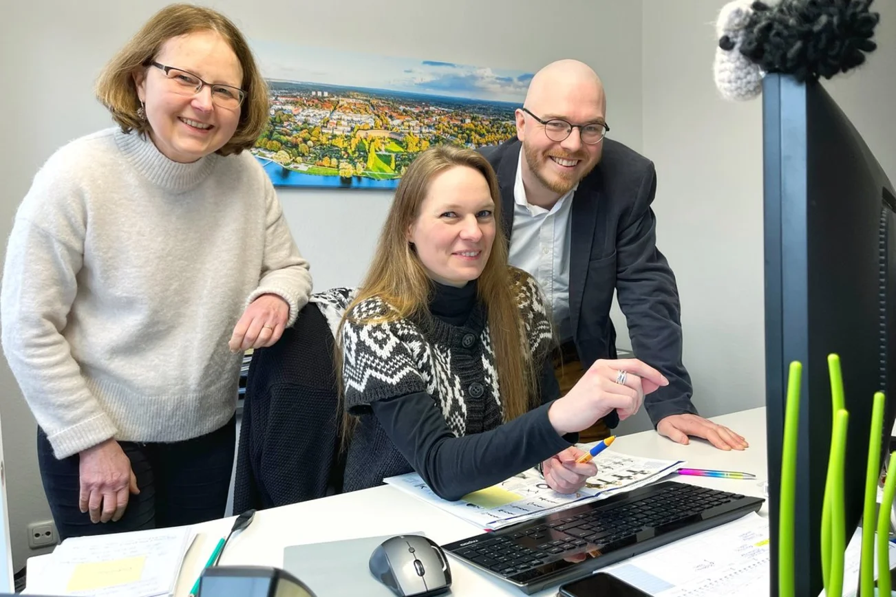 Drei Personen befinden sich in einem Raum und schauen auf einen Computer. 