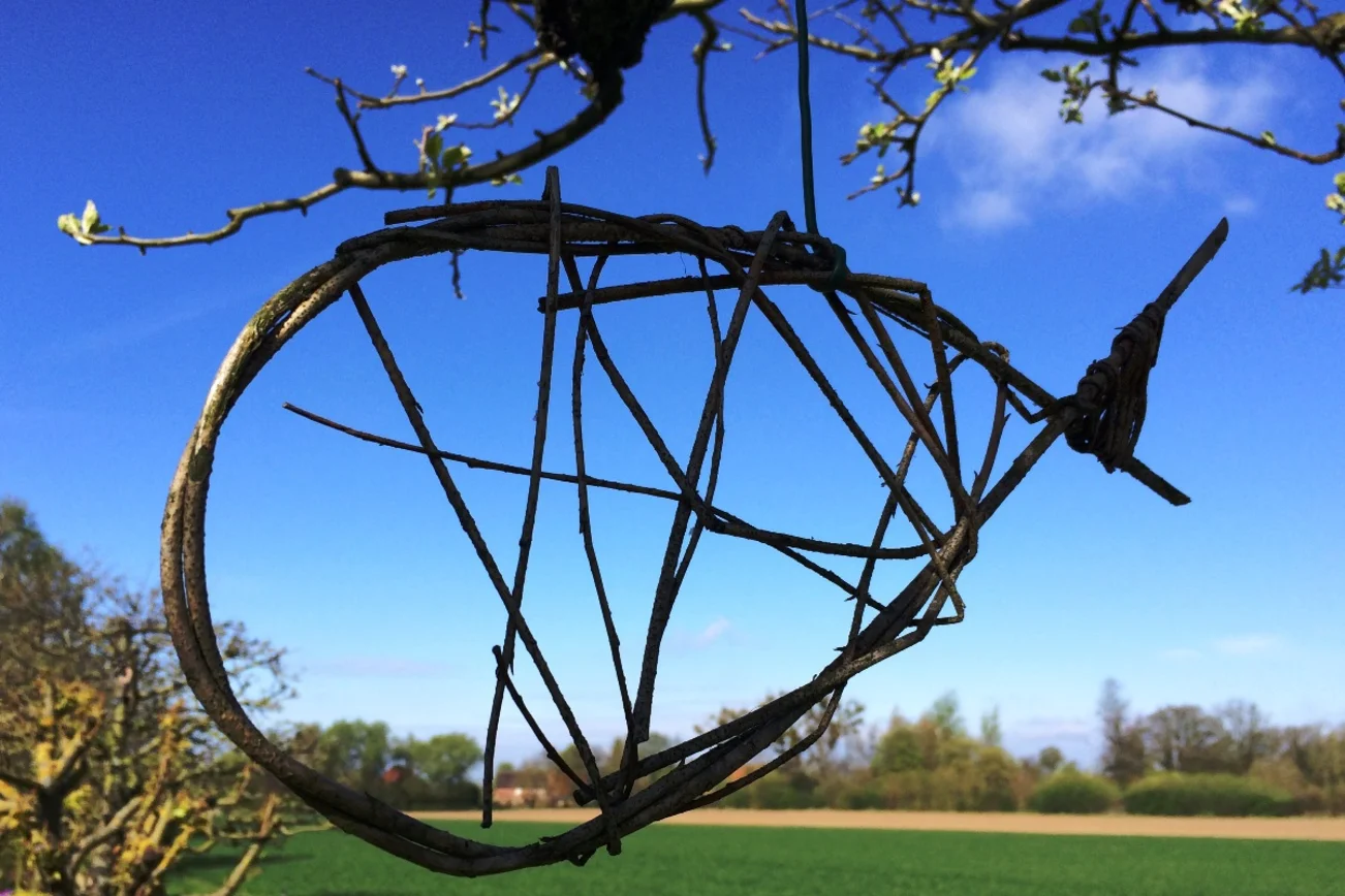 Die Umrisse eines Fisches aus Weidenruten. Sie hängen in einem Baum, im Hintergund Landschaft & Himmel.