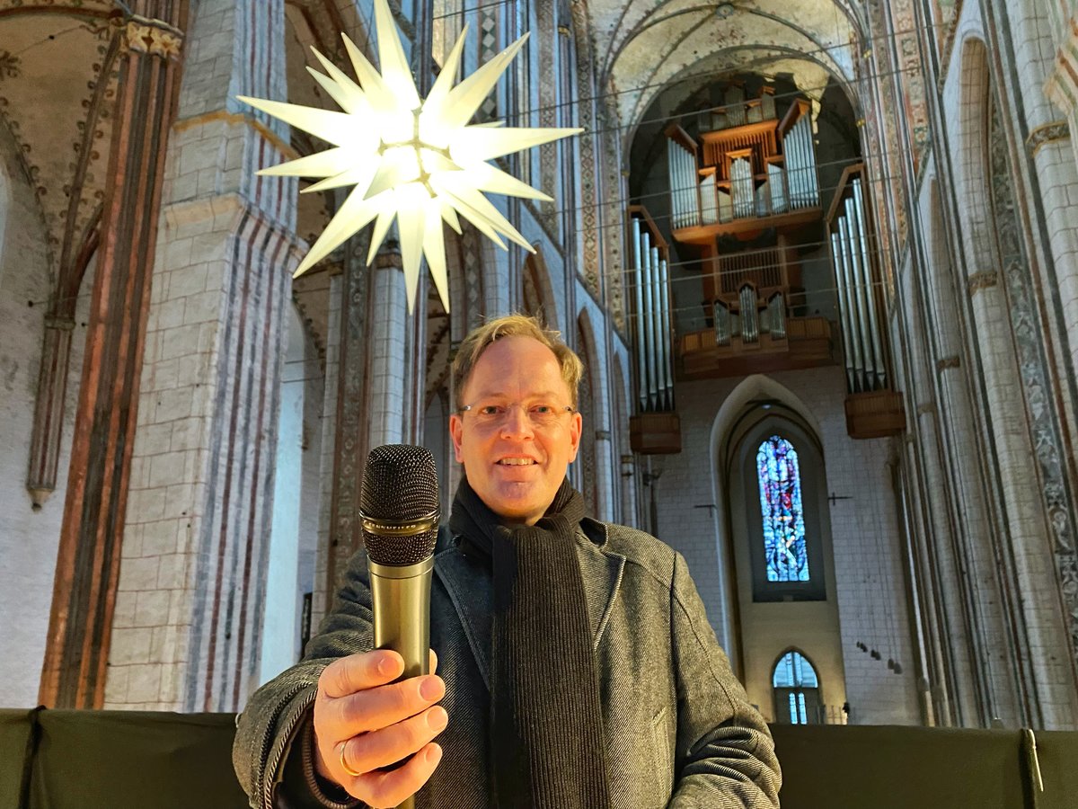 Mann steht in einer Kirche vor einem Leuchtstern und hält lächelnd ein Mikrofon in die Kamera