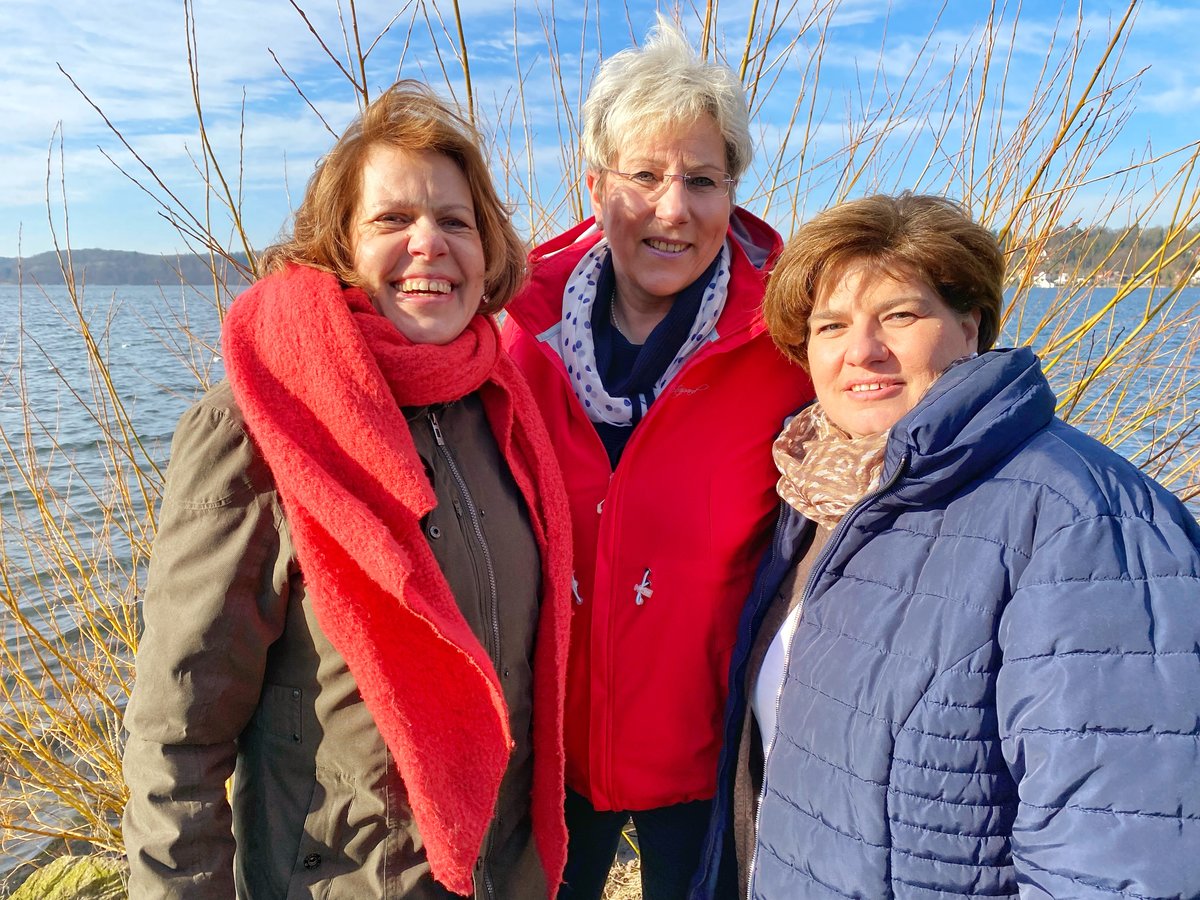 Drei Frauen stehen lächelnd am Rand eines Gewässers. 