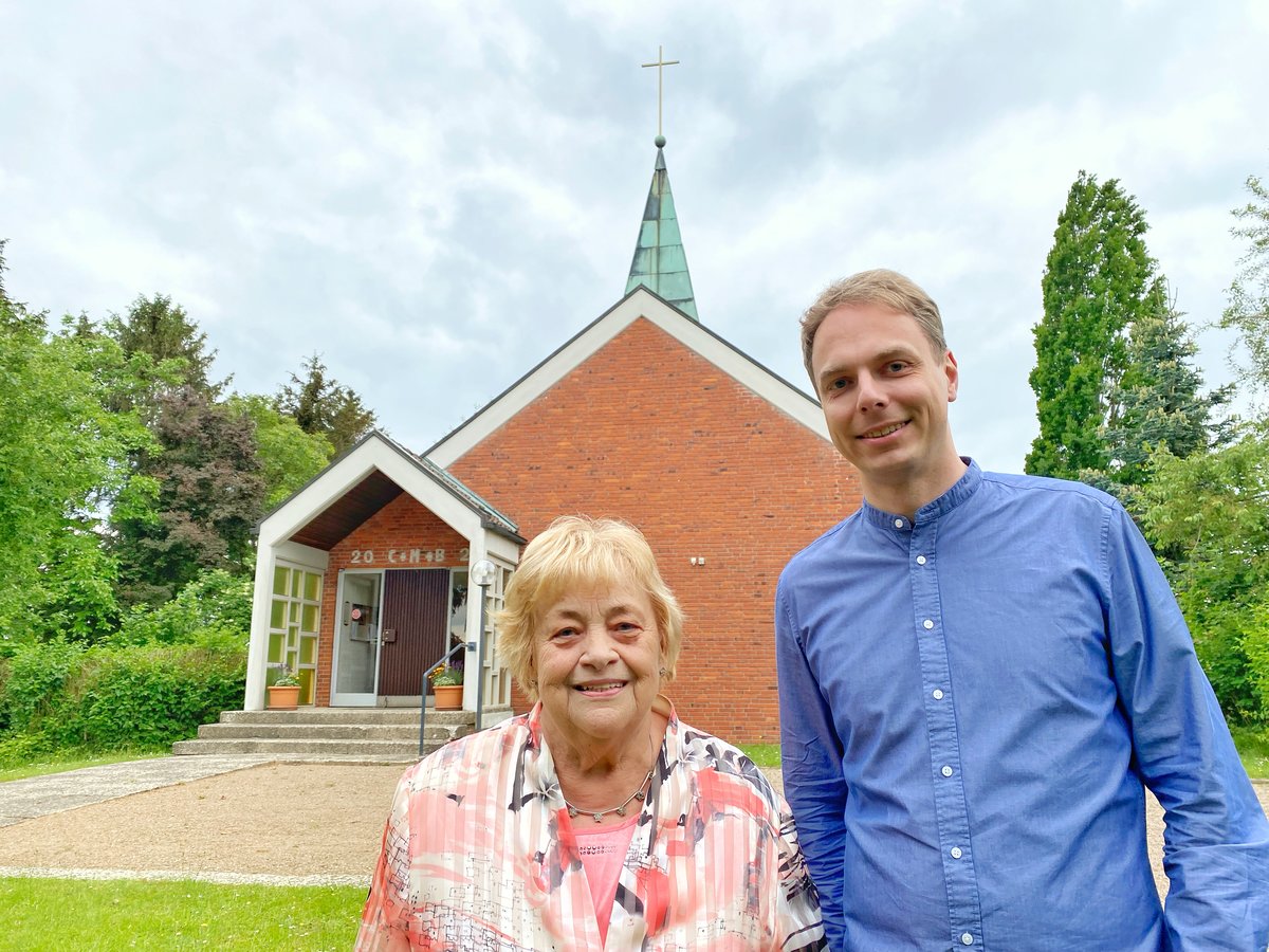 Zwei Personen stehen vor einer Kapelle und lächeln in die Kamera.