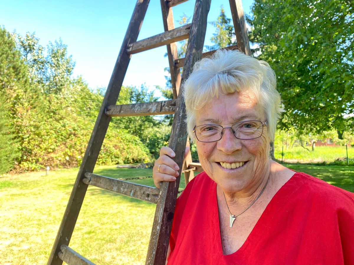 Eine Frau steht lächelnd an einer Leiter in ihrem Garten. 