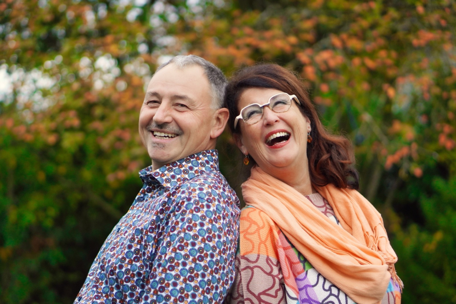 Ein Mann und eine Frau mit Brille stehen Rücken an Rücken in einem herbslichen Park und lachen in die Kamera.