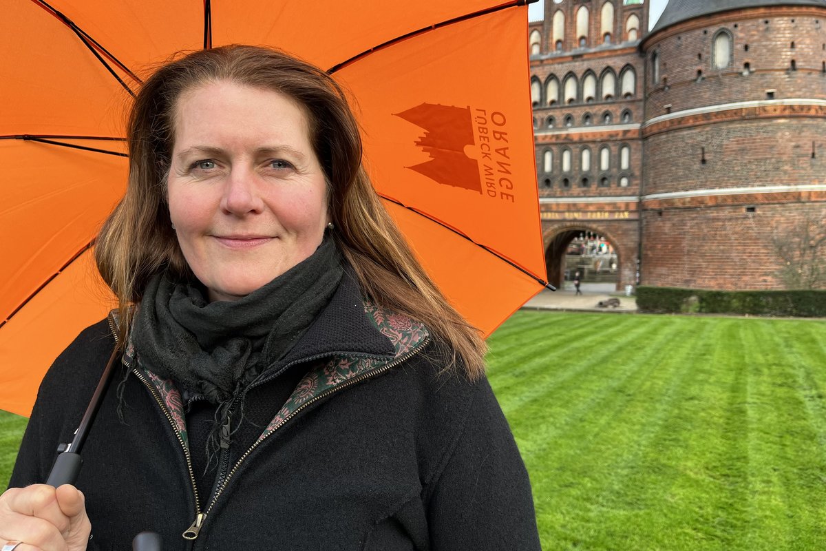 Eine Frau langen Haaren steht vor dem Lübecker Holstentor hält einen orangefarbenen Schirm.