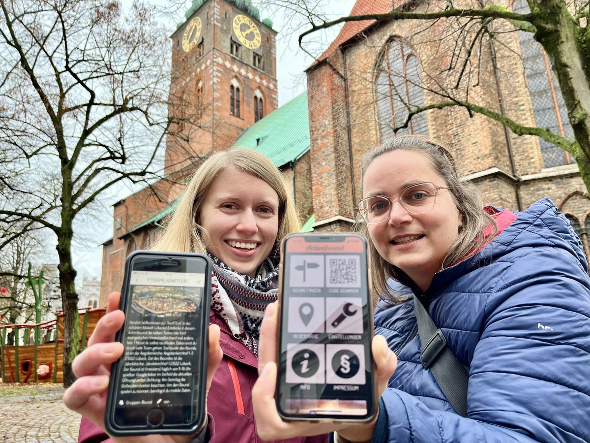 Zwei Frauen stehen vor einer Kirche und halten Smartphones in die Kamera. 