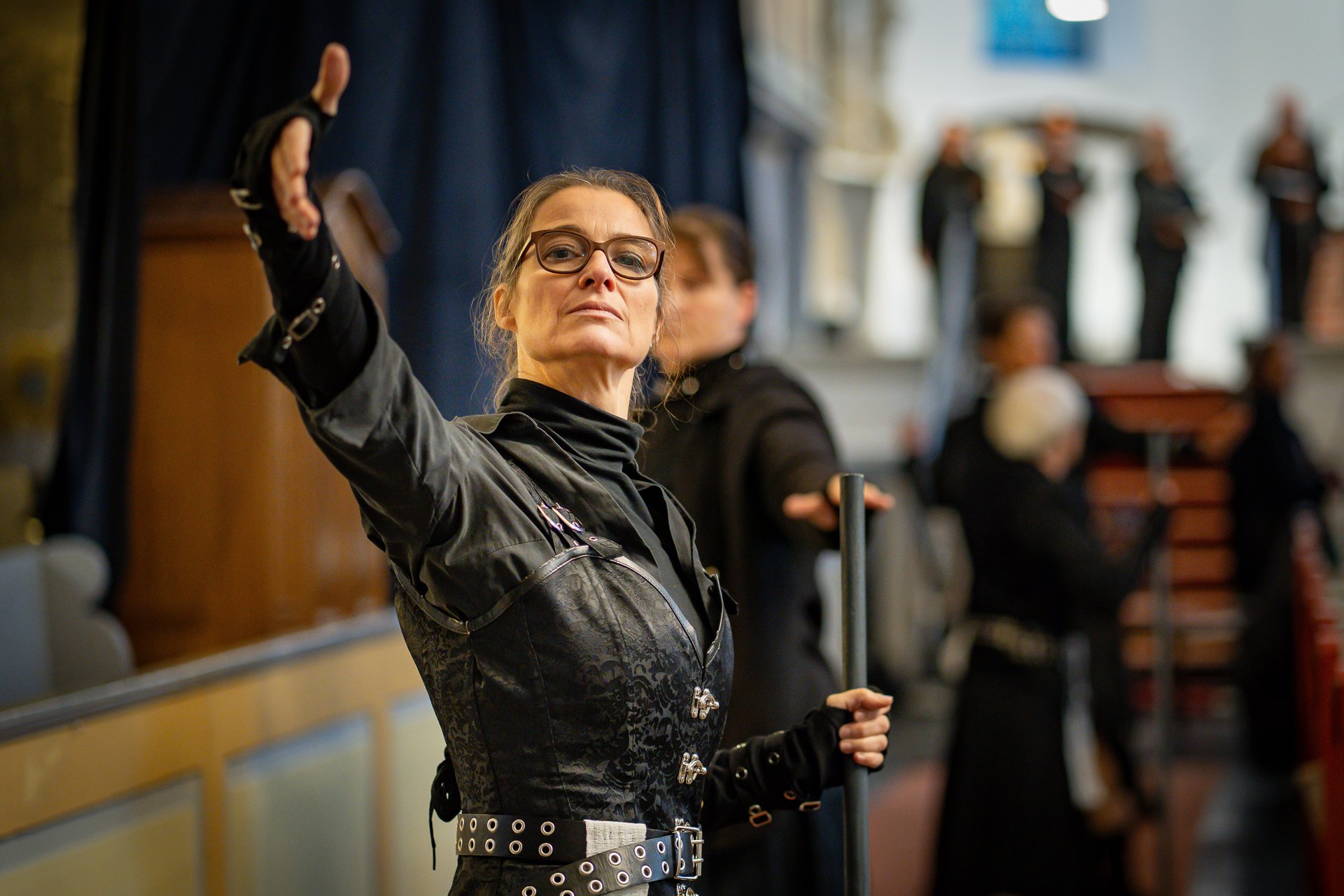 Eine Frau in schwarzer Kleidung steht in einer Kirche und macht eine ausladende theatralische Geste.