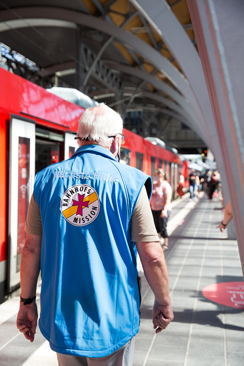 Ein Mitarbeiter der Bahnhofsmission geht ab Bahnsteig lang, auf der linken Seite sieht man einen roten stehenden Zug mit offenen Türen und die Rückseite der blauen Weste der Bahnhofsmission mit deren Logo