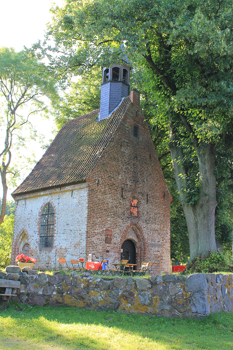 kleine Wegekapelle steht unter einem großen Baum mit vielen grünen Blättern, Vor der Kapelle stehen Klappstühle; im Vordergrund gibt es eine kleine Steinmauer