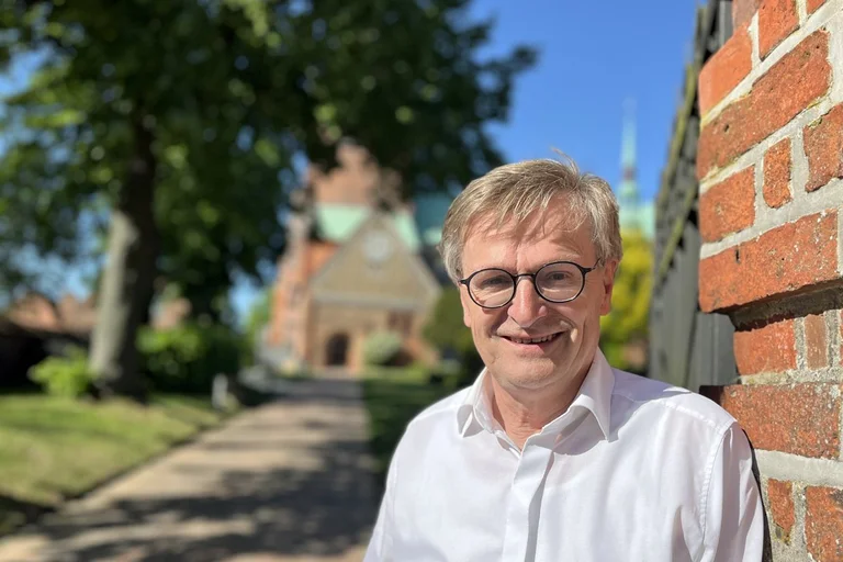 Ein Mann mit Brille steht an einer Backsteinmauer und lächelt in die Kamera.