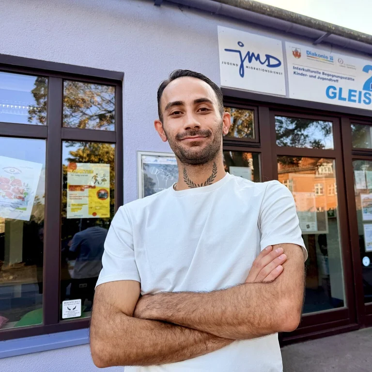 Ein Mann mit weißem T-Shirt und dunklen kurzen Haaren und Drei-Tage-Bart steht vor einem Gebäude mit großen Türen und Fenstern. Er hat die Arme vor sich verschränkt.