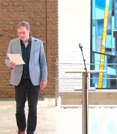 Ein Mann hält einen Zettel in der Hand auf den er schaut, rechts neben ist steht ein Mikrophon und ein Stehpult, weiter rechts ein großes Holzkreuz und ganz im Hintergrund ist ein buntes Kirchenfenster