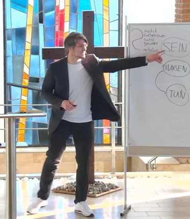 Ein Mann steht vor einem Holzkreuz und einer Tafel und zeigt mit dem Finger auf diese