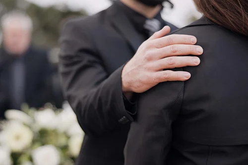 Im Hintergrund verschwommen sieht man einen weißen Blumenstrauß und eine Person mit dunklem Anzug. Im Vordergrund scharf abgebildet hält eine Mann, dessen Kopf man nicht sehen kann, seine rechte Hand tröstend auf der Schulter einer Frau in schwarzer Kleidung und dunklen Haaren. Auch ihr Gesicht kann man nicht sehen. Der Mann trägt ebenfalls schwarze Kleidung: Ein schwarzes Sakko und ein dunkles Hemd mit dunkler Krawatte.