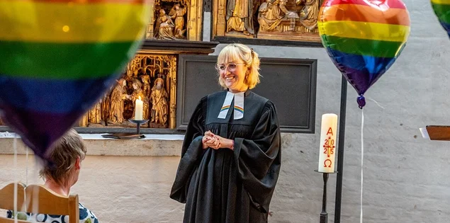 Eine Frau im Talar steht in einer Kirche mit bunt-gestreiften Helium-Herz-Luftballons