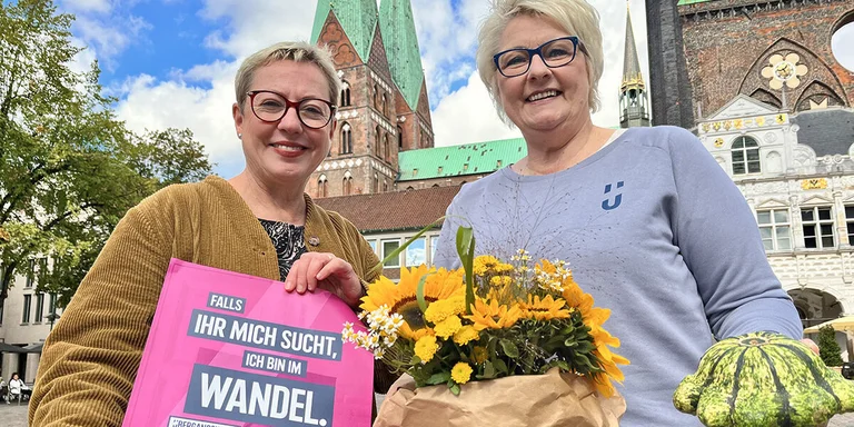 Zwei Frauen mit Blumen und Kürbis in den Händen lächeln auf dem Lübecker Marktplatz in die Kamera.