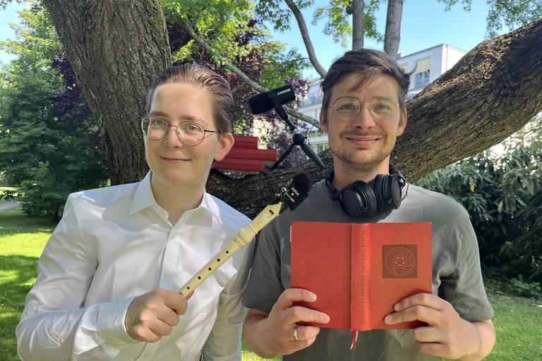 Zwei Männer stehen vor einem Baum und halten eine Blockflöte und ein rotes Gesangbuch.