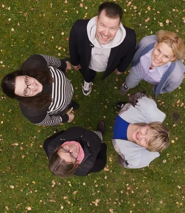 Eine Gruppe Menschen steht im Kreis und schaut nach oben und wird von einer Drohne fotografiert. 