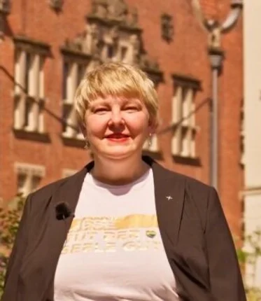Eine Frau mit kurzen blonden Haaren steht zwischen Pflanzen in einer Altstadt.