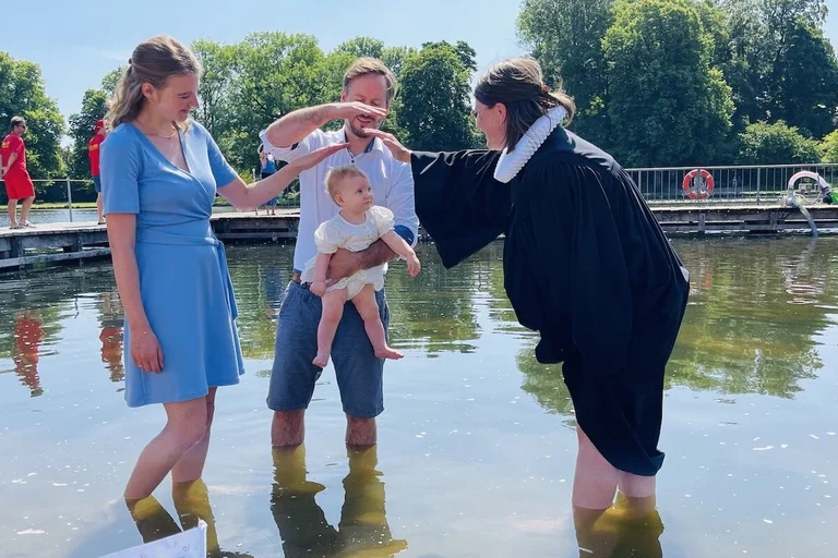Eine Pastorin tauft ein kleines Mädchen im Krähenteich. Der Vater hält es hoch, die Mutter schaut zu freut sich. Beide Eltern halten ihre Hände als Segen über den Kopf des Kindes. 