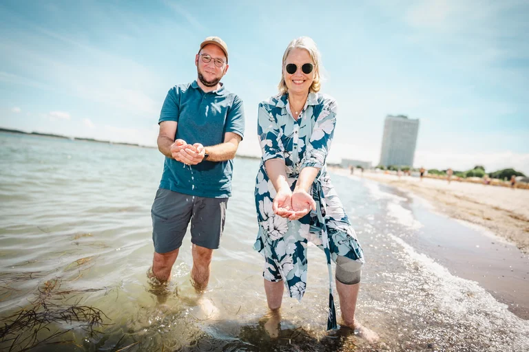 Zwei Personen stehen in der Ostsee, haben Wasser mit den Händen geschöpft; im Hintergrund ist das Travemünde Maritim zu sehen.
