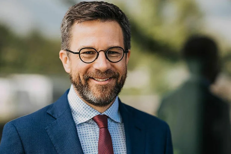 Das Portrait eines Mannes mit Brille, kurzen Haaren, Bart. Er trägt ein dunkles  Sakko.