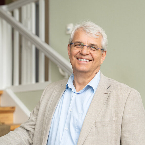 Ein Mann mit grauen Haaren und Brille schaut lächelnd in die Kamera. Er trägt ein hellblaues Hemd und ein helles Sakko.