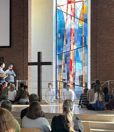 Mehrere Menschen in einer Kirche-2 Personen stehen hinter einem Kreuz in einem Taufbecken, andere stehen darum herum und einige auf Stühlen vor ihnen