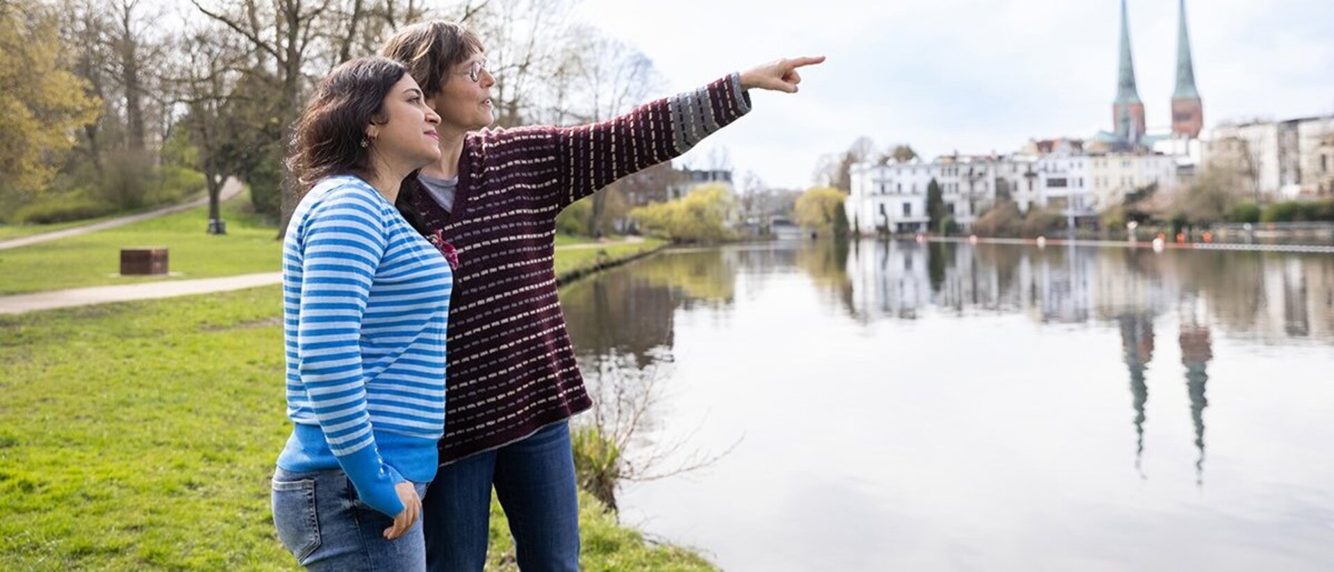 Zwei Frauen stehen nebeneinander an einem Gewässer. Die eine Frau zeigt in die Ferne. Im Hintergrund ist die Stadt Lübeck zu sehen mit den Türmen von St. Marien und dem Petriturm.